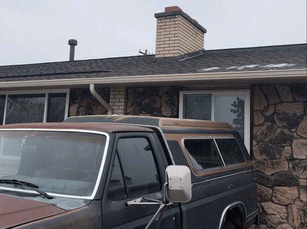 A weathered vintage car parked in front of a stone-faced house with a chimney.