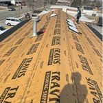 A worker on a roof under construction with wooden beams and protective paper. Shadow of the photographer visible.