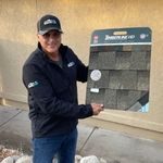 Person in a black jacket presenting a roofing shingle sample board outdoors.